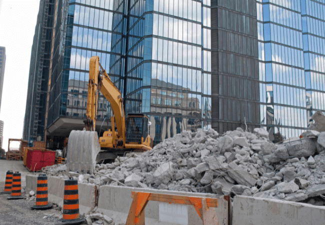 Excavator breaking down concrete rubble in an urban demolition site, surrounded by high-rise buildings
