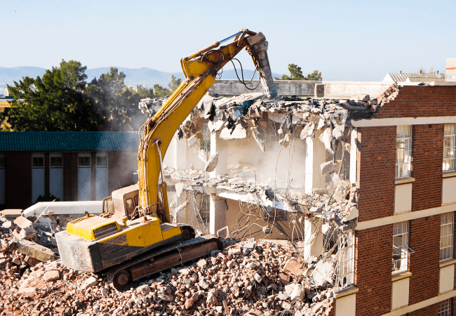 Excavator tearing down the top floor of a building, with debris and rubble scattered around the demolition site